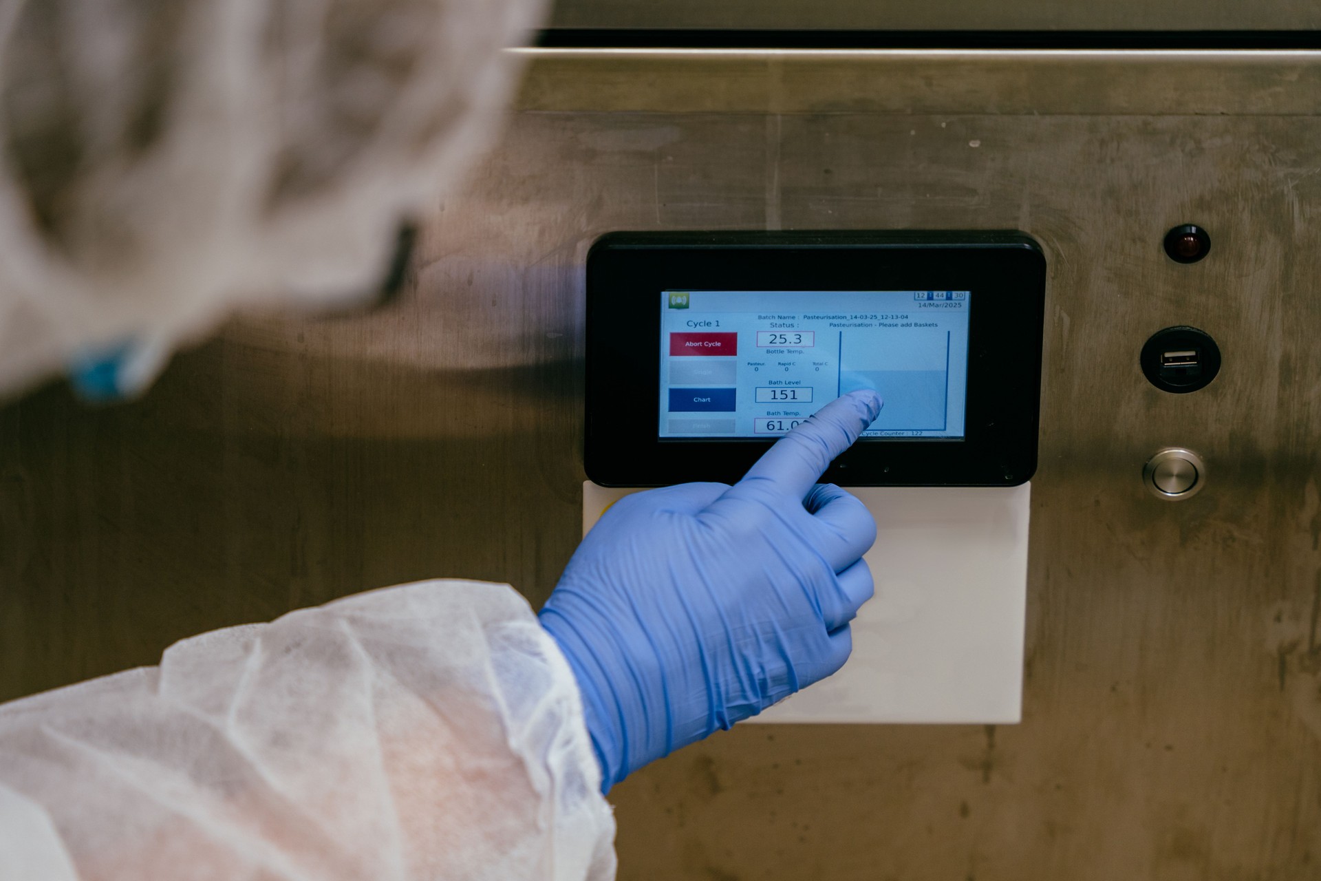 Close-up of gloved hand adjusting settings on pasteurizer control panel at milk bank lab during thermal treatment.